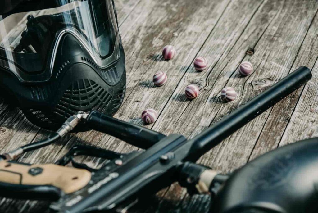 Paintball-Maske, Marker und mehrere lila-weiß gestreifte Paintballs auf Holzboden.
